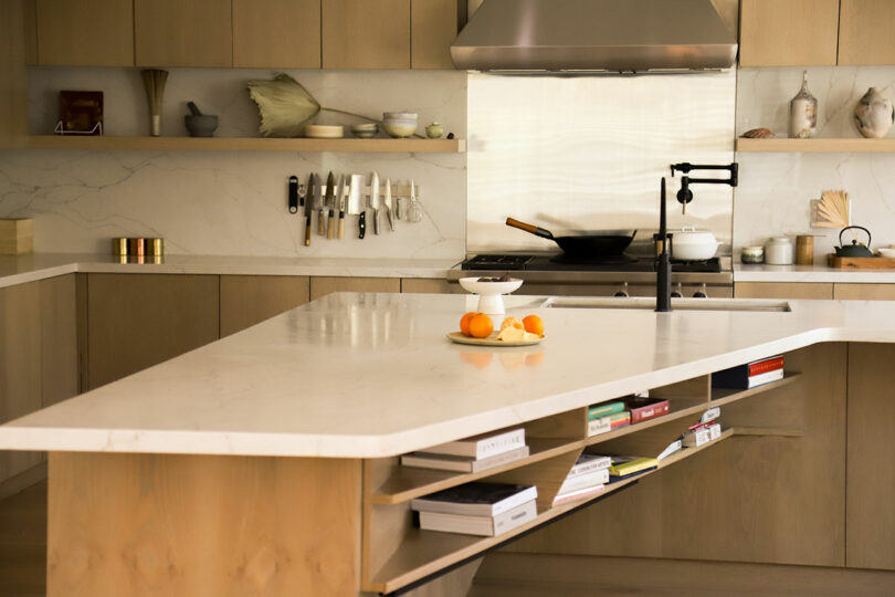 The modern kitchen, with its marble island adorned with fresh oranges and a bowl, reflects Amanda Gunawan's design ethos. Knives neatly line the back wall, complementing the stovetop hood and elegant wooden cabinetry.