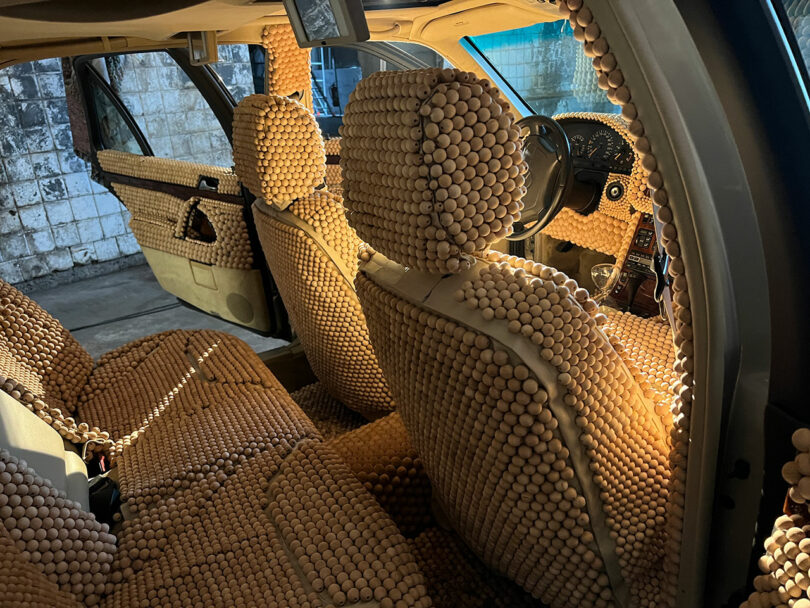 Car interior covered entirely in wooden bead seat covers, including seats, doors, and dashboard. Sunlight casts a warm glow on the beads.