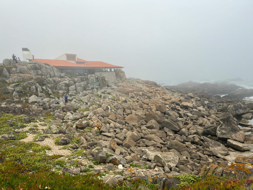 Foggy coastal scene with rocky terrain and a building with a red roof in the background.