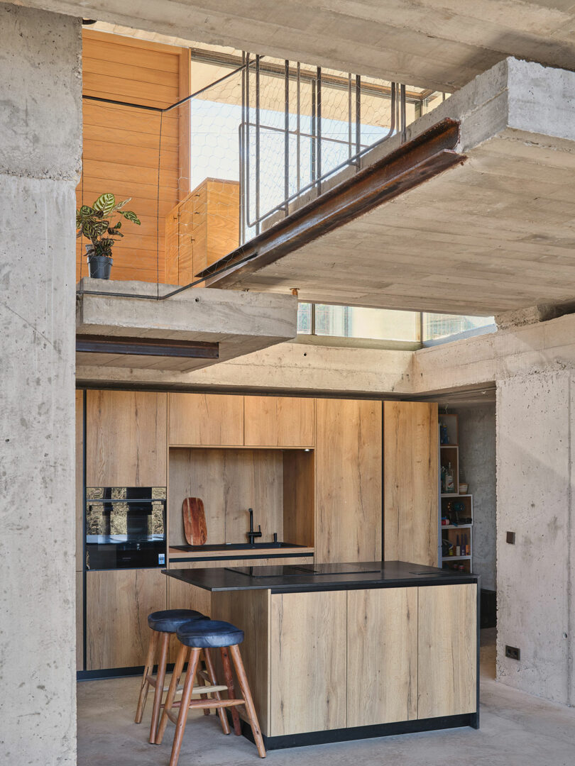 Modern kitchen with wood cabinets and black countertops, featuring a small island with two stools. Exposed concrete walls and ceiling, with a staircase leading to a mezzanine.