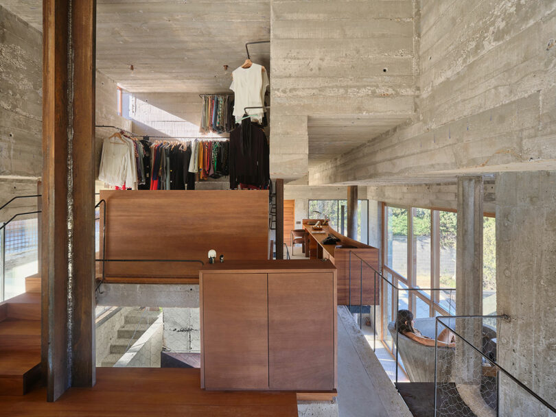 Concrete interior with multi-level design, featuring wooden furnishings. A rack displaying clothes is visible above. Large windows provide ample natural light. A person is sitting by the stairs.