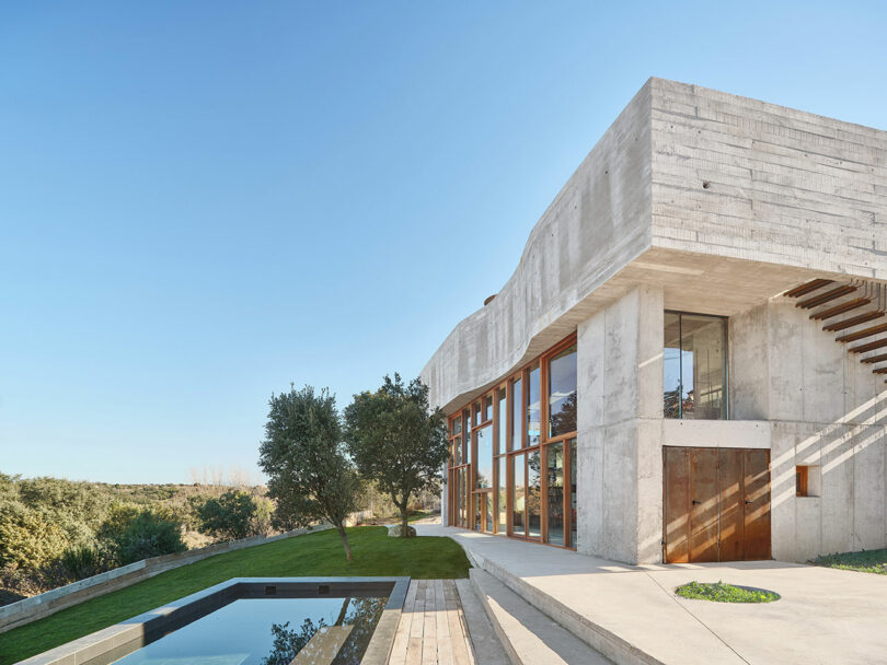 Modern concrete house with large windows, adjacent to a reflecting pool and landscaped lawn, set against a clear blue sky.