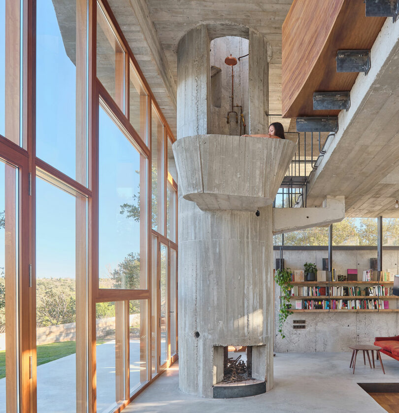 Concrete cylindrical structure inside a modern room with large windows, bookshelves, and greenery outside; person peering from inside the upper part of the structure.