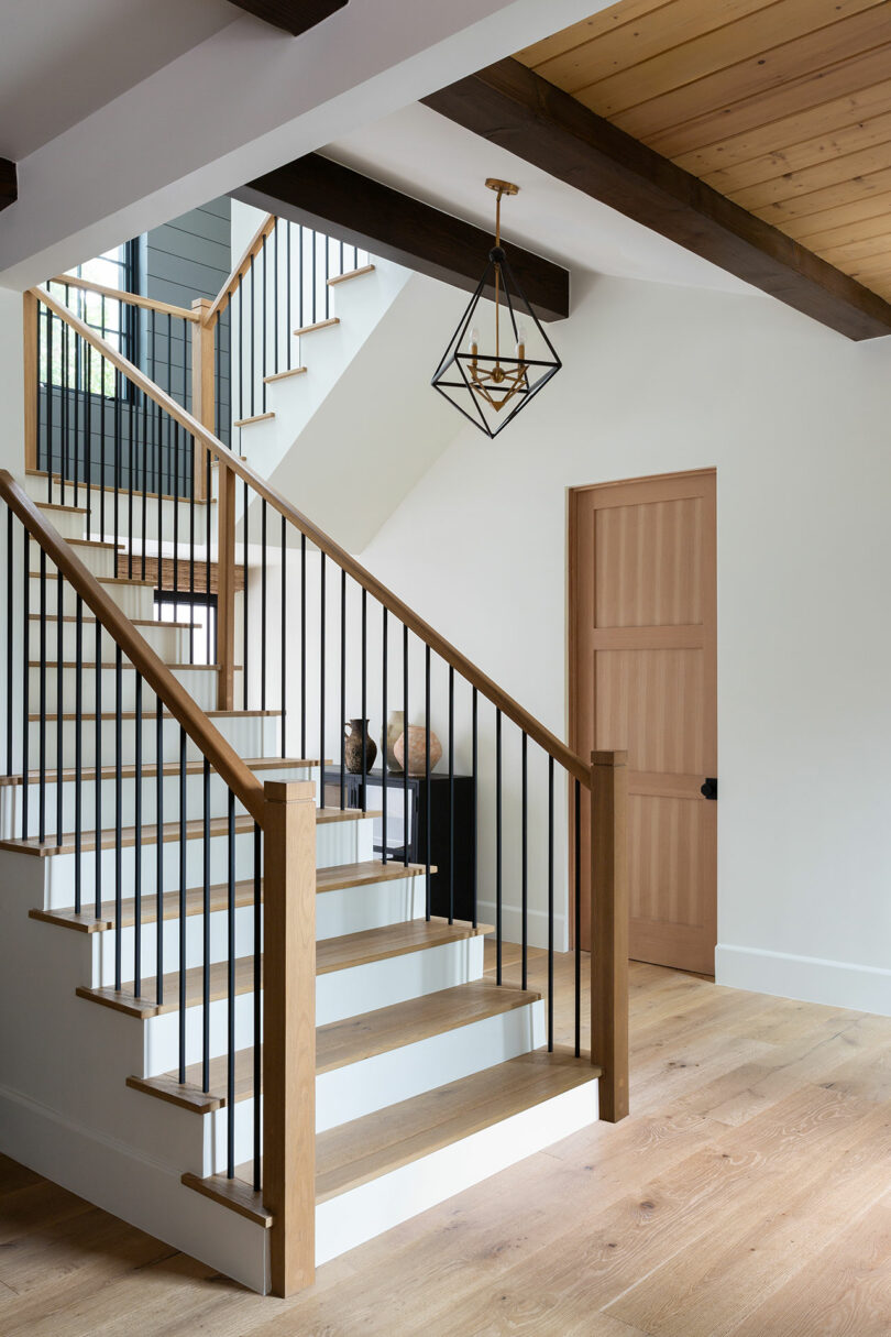 Modern staircase with wooden steps and black railings, pendant light fixture above, and wooden door on the right in a bright, airy hallway.