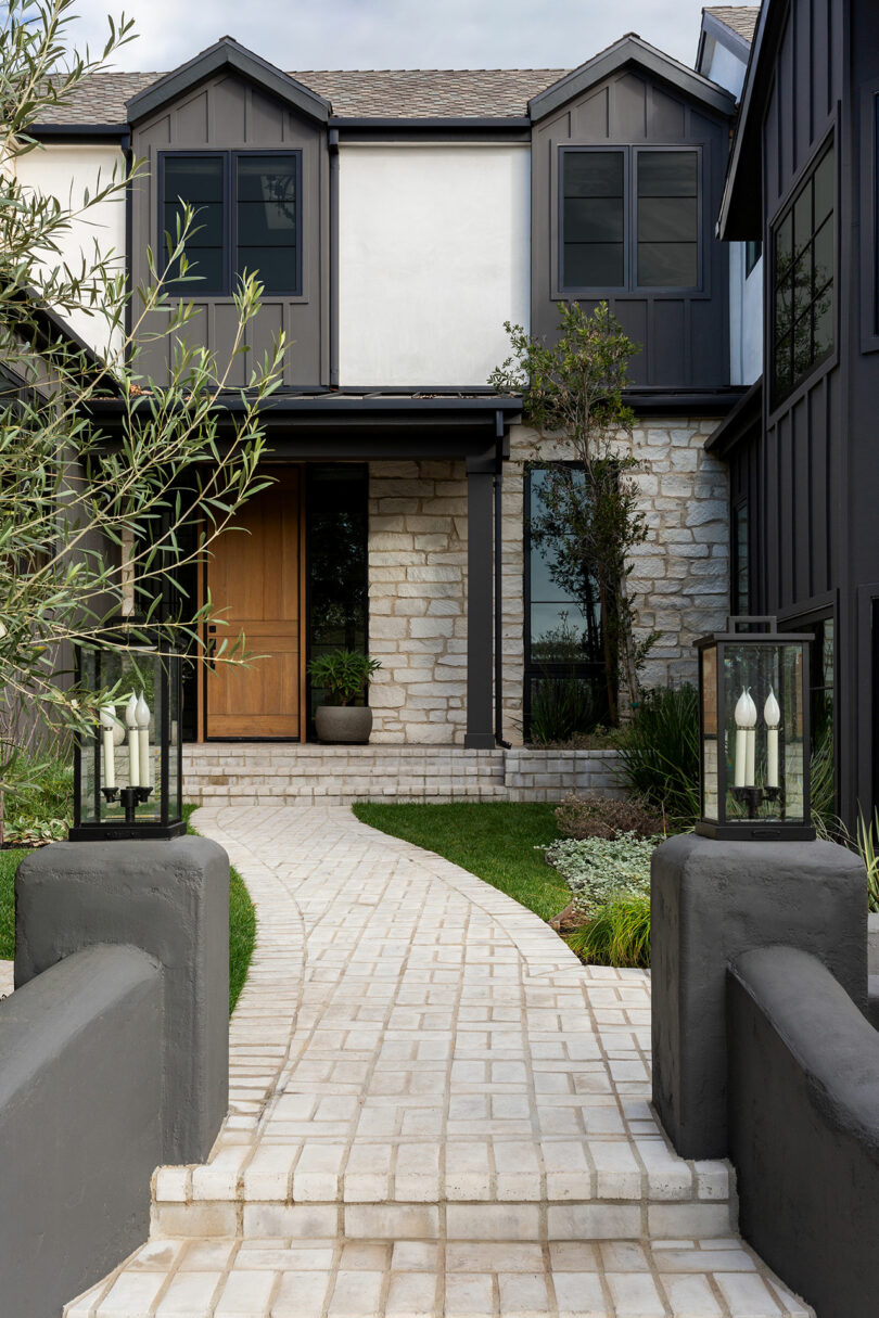 Modern house exterior with dark accents, a wooden door, and stone facade. Pathway leads through landscaped garden with small trees and plants. Two lantern-style lights flank the entrance.