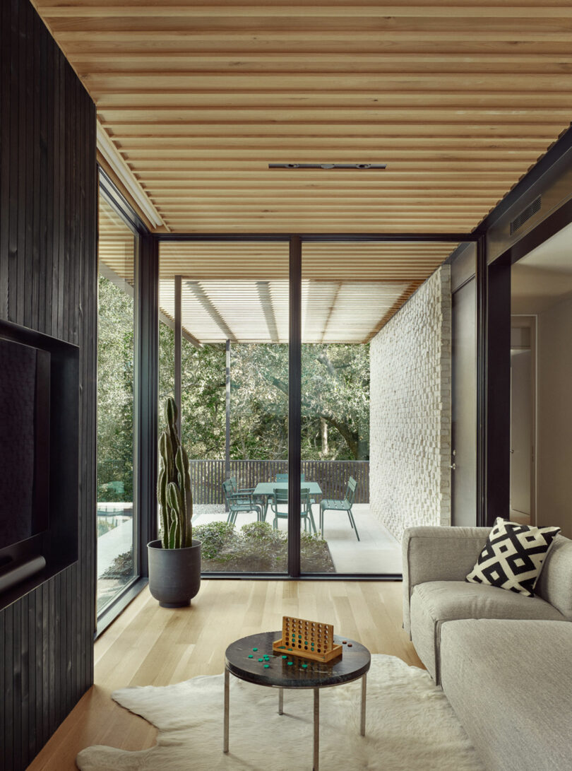 Modern living room with large glass doors, wooden ceiling, and light-colored flooring. A gray sofa with a geometric pillow is next to a round table with a small game on it. A patio is visible outside.