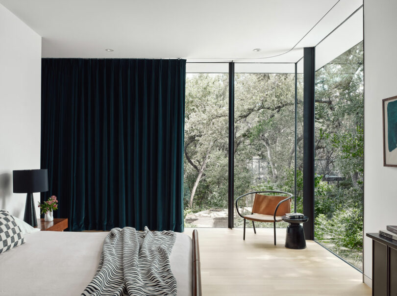 Modern bedroom with large windows showcasing a forest view. Features a bed with a striped blanket, black curtain, and a seating area with a chair and small table. Minimalist decor.