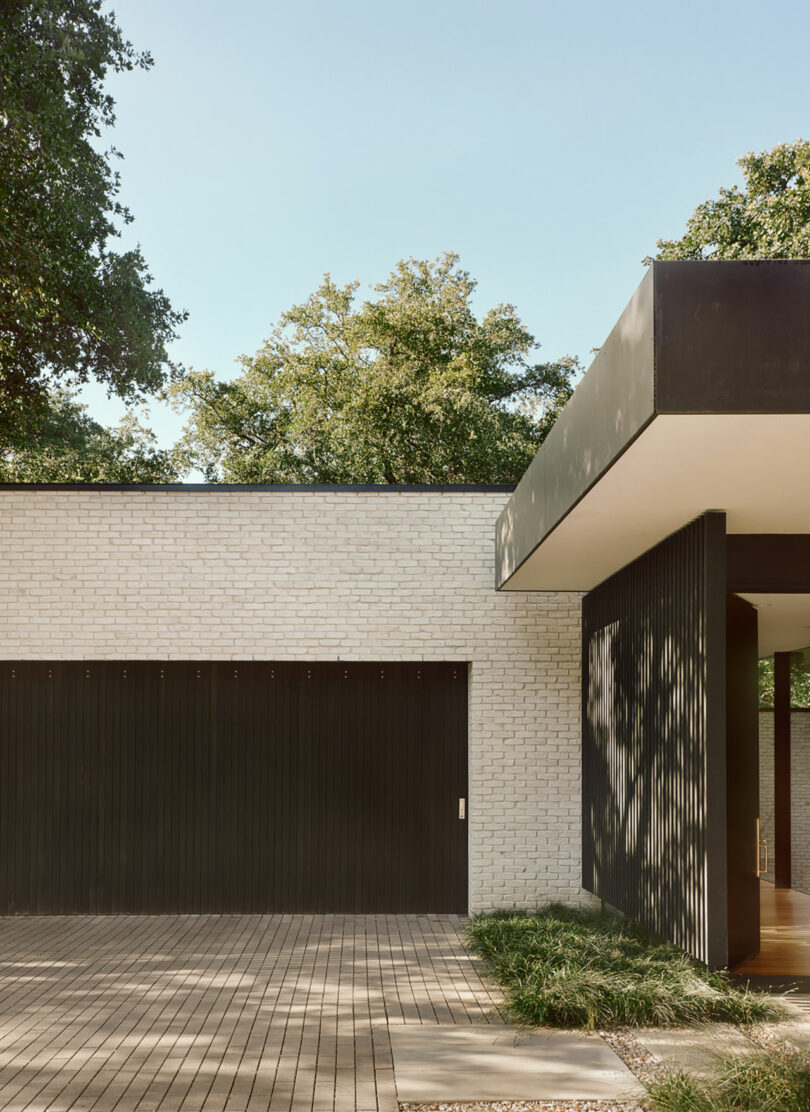 Modern house exterior with white brick and black accents, featuring a large garage door, flat roof, and paved driveway framed by grass, under clear blue sky and surrounded by trees.