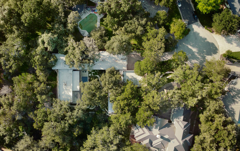 Aerial view of a residential area with tree-covered houses and a small swimming pool, surrounded by greenery and winding roads.