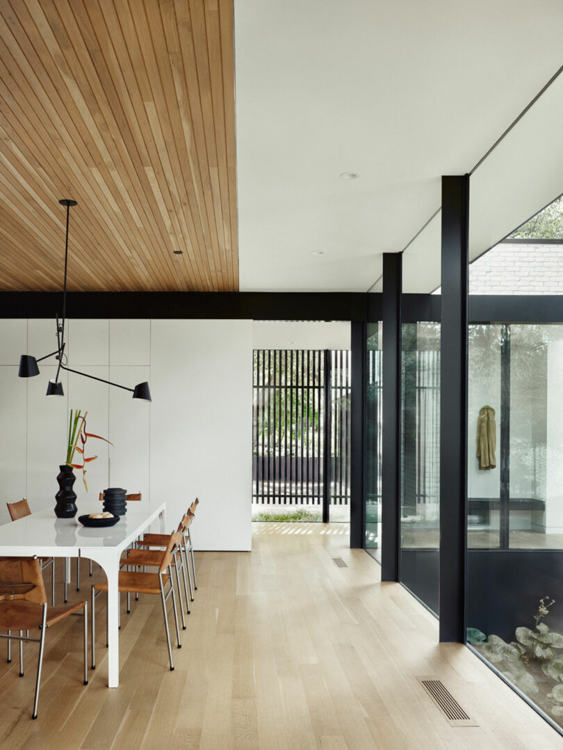 Modern dining area with a white table, wooden chairs, black pendant light, and a wood-paneled ceiling. Large windows and a decorative slatted screen offer views of greenery outside.