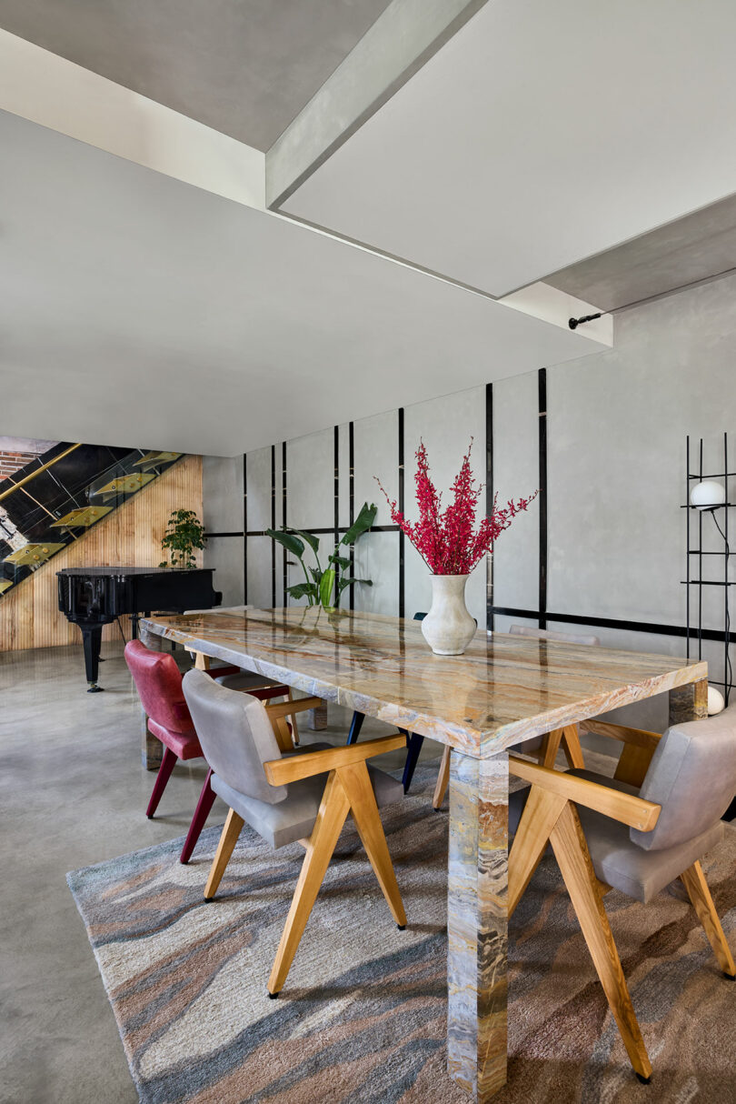 Modern dining room with a large marble table, surrounded by six chairs in red and gray. A vase with red flowers sits on the table. A grand piano is visible in the background near a staircase.
