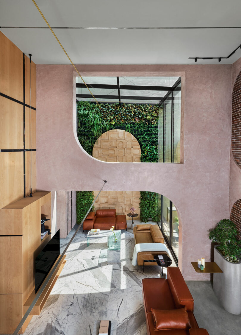 Modern living room with high ceilings, brown leather sofas, a glass coffee table, and large windows showing green plants outside. Pink textured walls have arched cutouts revealing an upper level.