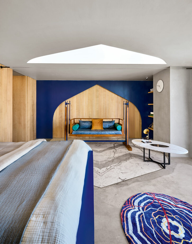 Modern bedroom with a blue and wood accent wall featuring a built-in bench. A bed with blue bedding, a patterned rug, and a marble-topped table are present under a uniquely shaped ceiling cutout.