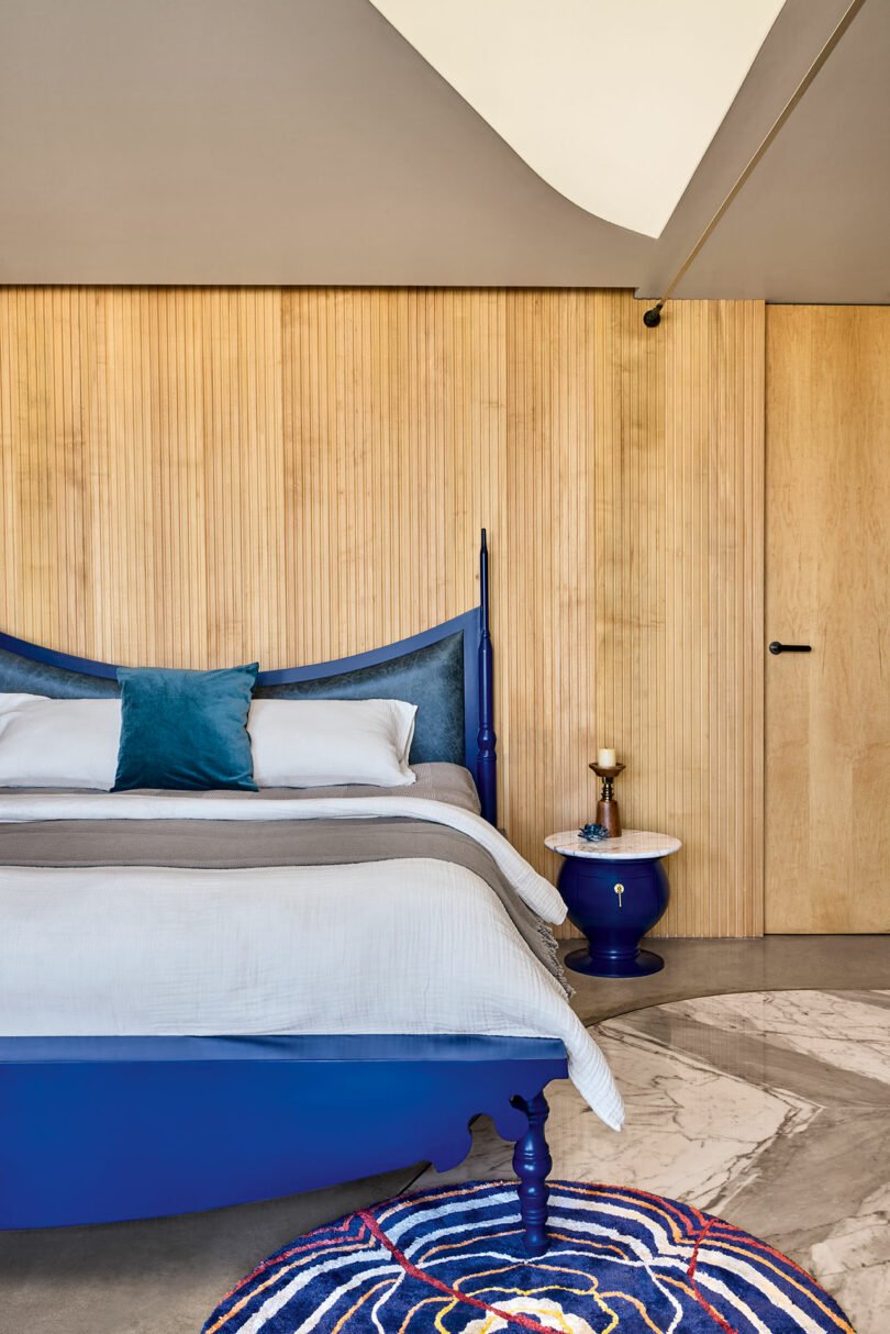 Bedroom with a blue bed, a teal pillow, a small side table with a lamp, wooden panel walls, and a patterned rug on the floor.