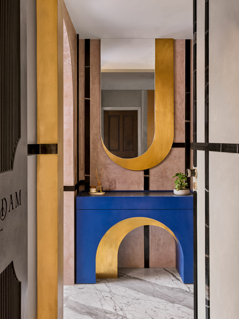 Modern hallway with pink walls, a blue console table featuring a gold arch design, and a large U-shaped gold mirror. Marble floor and a small plant on the table complete the decor.