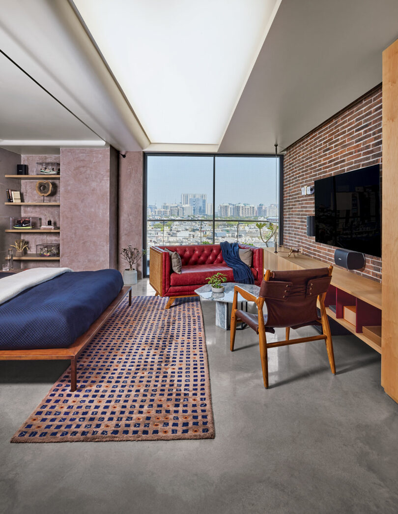 Modern bedroom with city view, featuring a bed with a blue cover, a red sofa, a brown chair, a wall-mounted TV, and a patterned rug on a polished concrete floor.