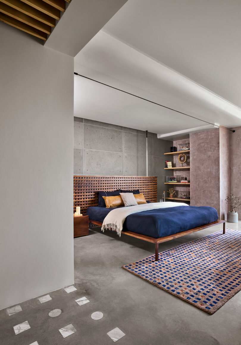 Modern bedroom with a concrete wall, wooden headboard, and navy bedding. Shelves with decor are on the left, a plush rug on the floor, and a small bedside table with a candle on the right.