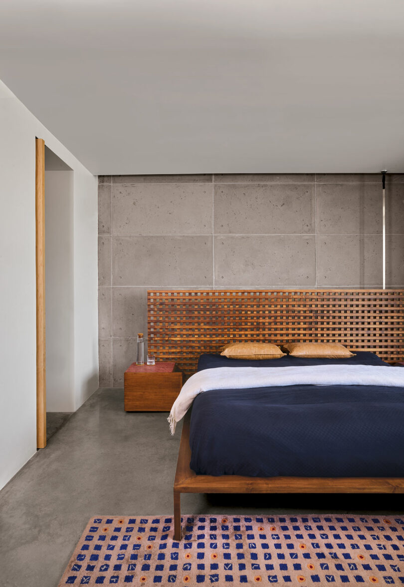 Modern bedroom with a concrete accent wall, wooden headboard, and navy bedding. There's a wooden nightstand with a bottle and glass. A colorful rug is on the concrete floor.