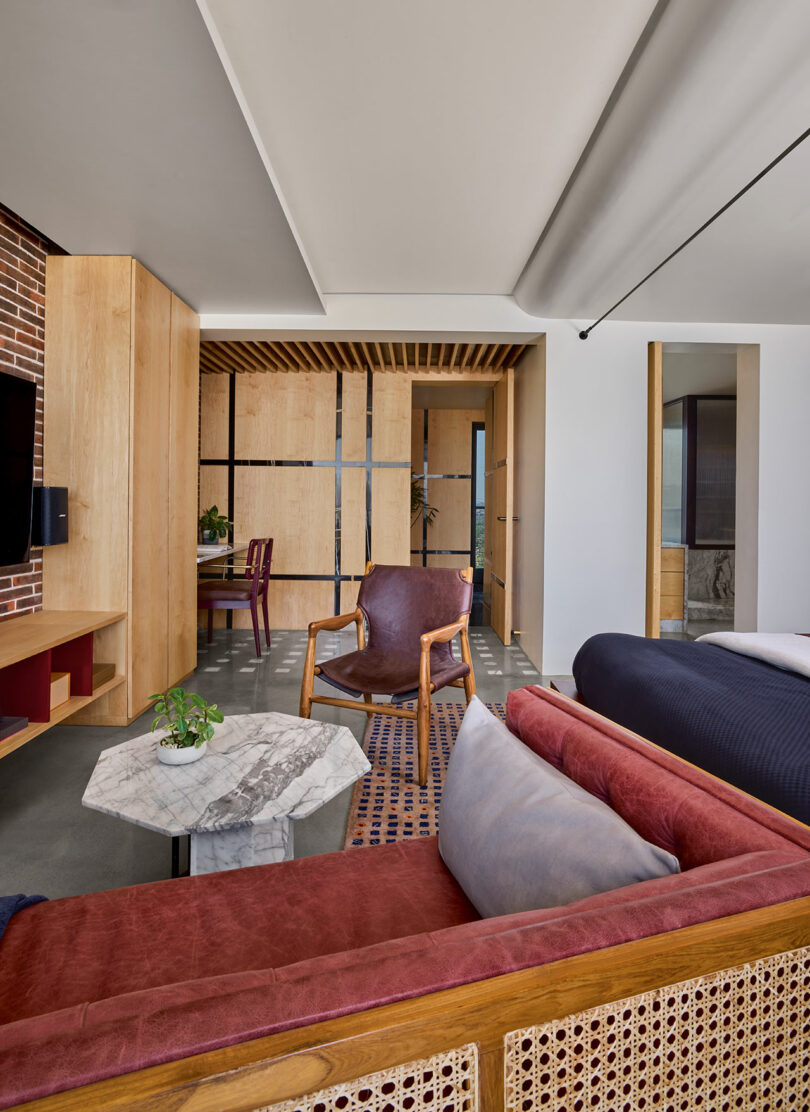 Modern living room with brick and wood accents, featuring a red couch, armchair, marble coffee table, and plant. Dining area with purple chair and wooden ceiling visible in the background.