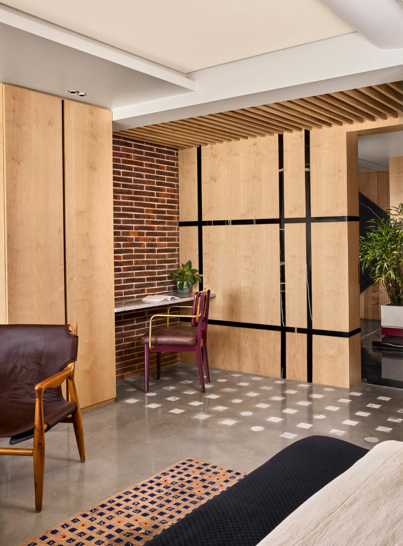 Modern interior with wood panels, exposed brick, and a small desk with a purple chair. Brown leather chair in the foreground and potted plant next to a partition wall. Geometric rug on floor.