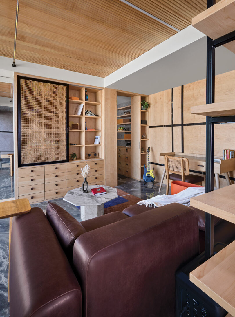 Modern living room with a brown leather sofa, marble coffee table, wooden shelving, and a textured wall panel. Room features a mix of open shelving, closed cabinets, and a light wood ceiling.