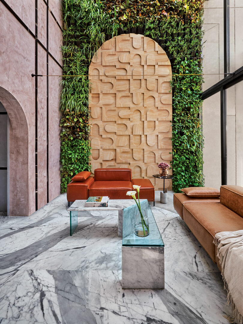Modern lobby with marble floor, brown leather sofas, glass coffee table, and vertical garden. Wooden artistic panel centered on a green wall, combining natural and contemporary design elements.