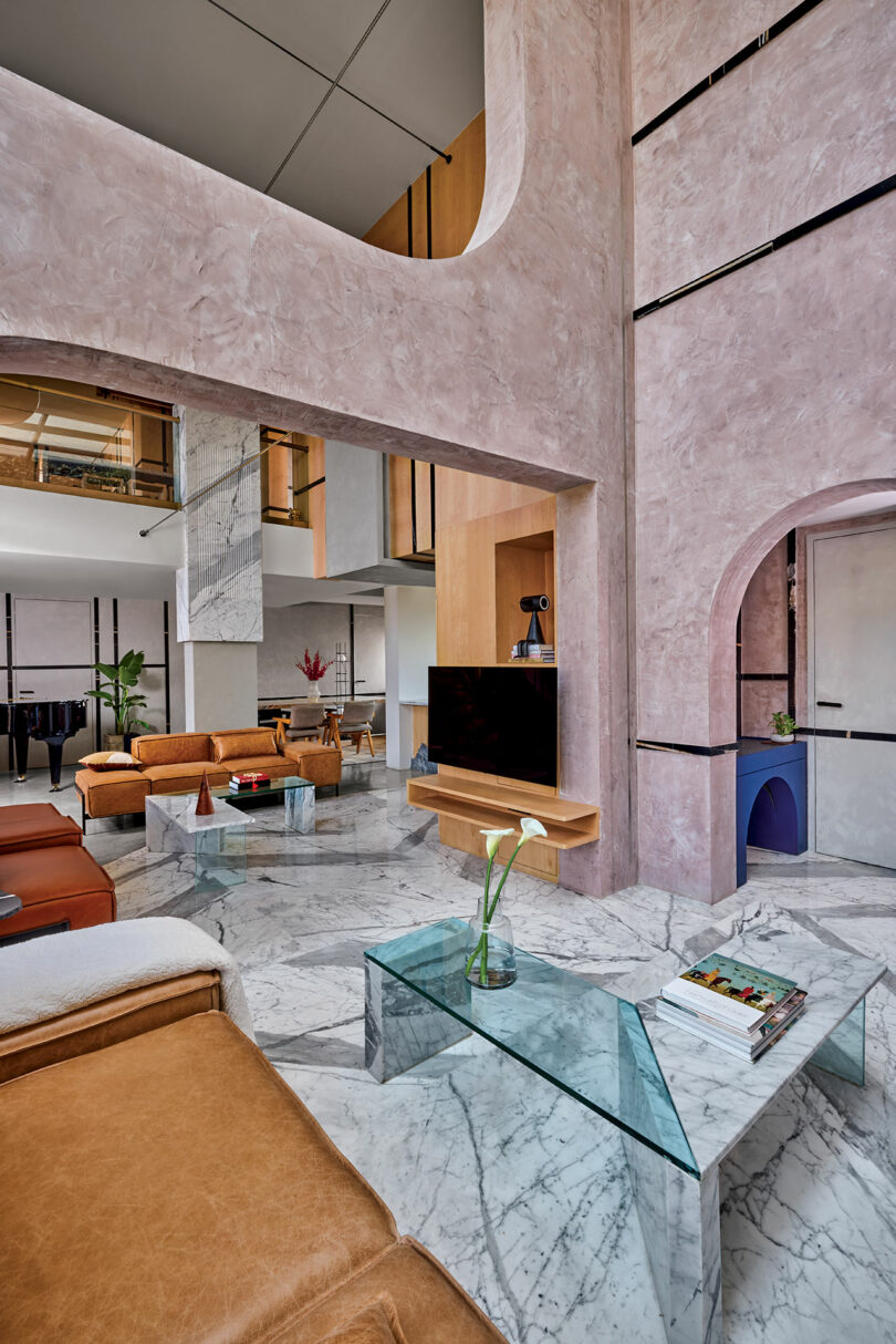 Modern living room with high ceilings, pink textured walls, marble floors, brown leather furniture, glass coffee table, and a TV. Archways and vases with flowers add decorative elements.