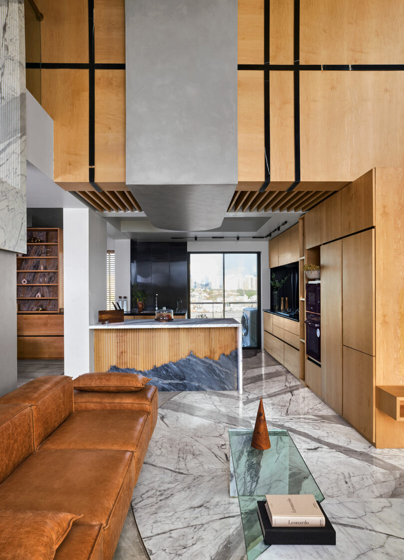 Modern living space with brown leather couch, marble floor, and a glass coffee table. Wood paneling and black accents highlight the kitchen area with city view through a large window.
