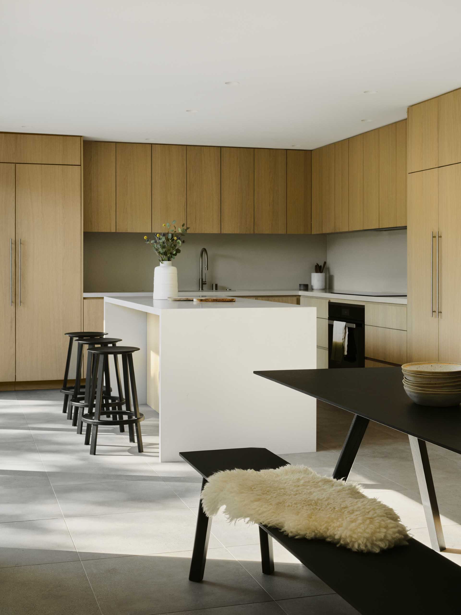A modern kitchen with wood cabinets and an island, opens up to the deck through a sliding door.