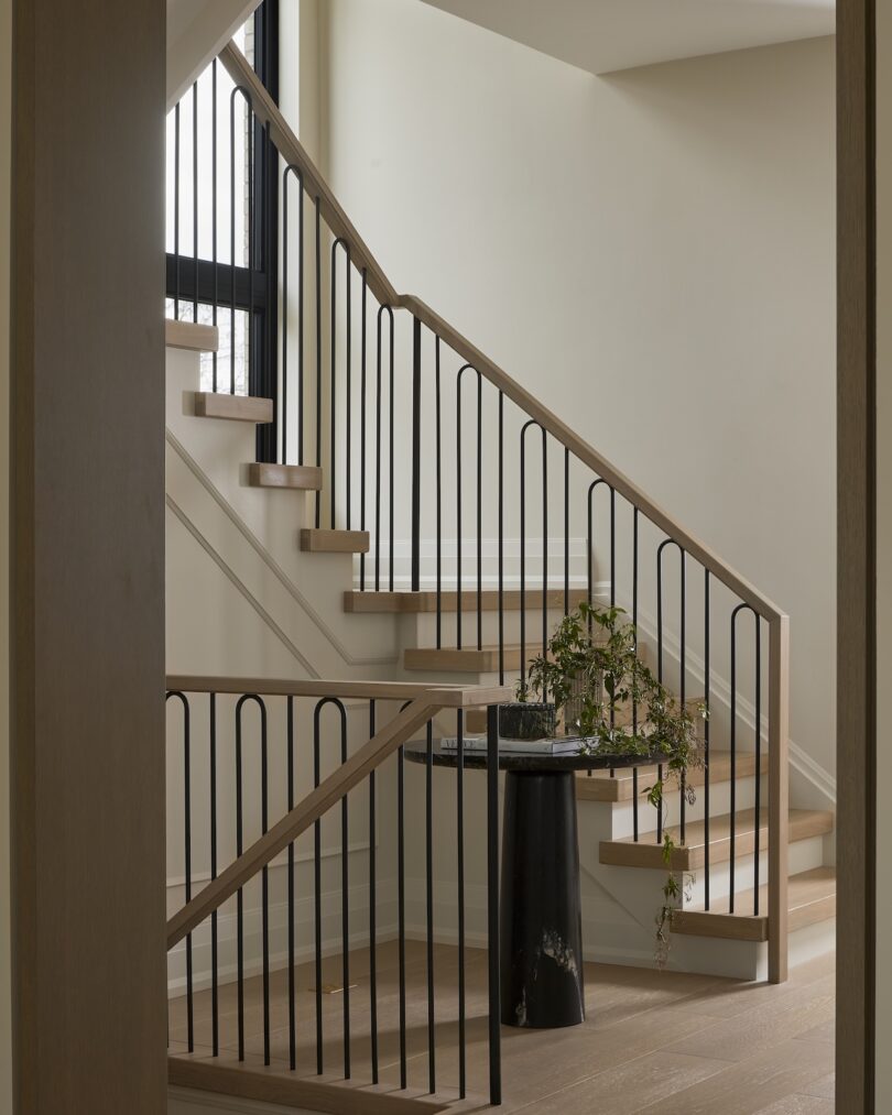Modern interior staircase with light wood steps and black metal railings. A small black pedestal with a potted plant sits at the foot of the stairs. Natural light filters through a window.