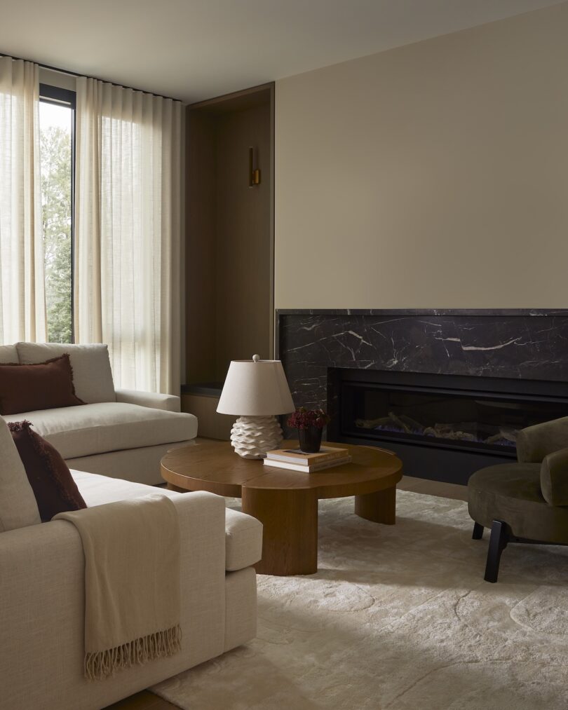 A modern living room with two sofas, a round wooden coffee table, a black marble fireplace, a small lamp, and sheer curtains covering tall windows.
