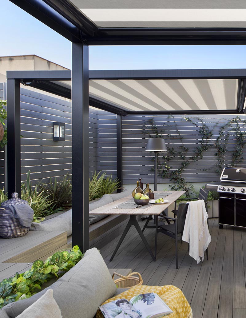 This sleek terrace features a beautifully designed outdoor dining area, with a long table flanked by stylish chairs on one side and a built-in banquette on the other. The arrangement creates a welcoming, intimate setting for meals, perfect for hosting family or friends. The minimalist design is complemented by lush greenery, enhancing the peaceful atmosphere. 