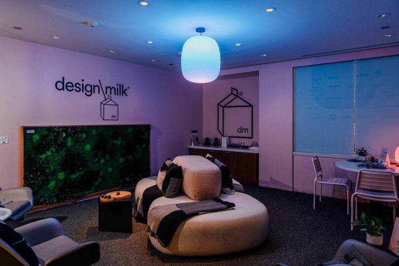 Modern Designer Wellness Lounge featuring a curved white sofa, chic chairs, wall art reading "design milk," a lush green plant panel, a round table with chairs, and a glowing ceiling light.