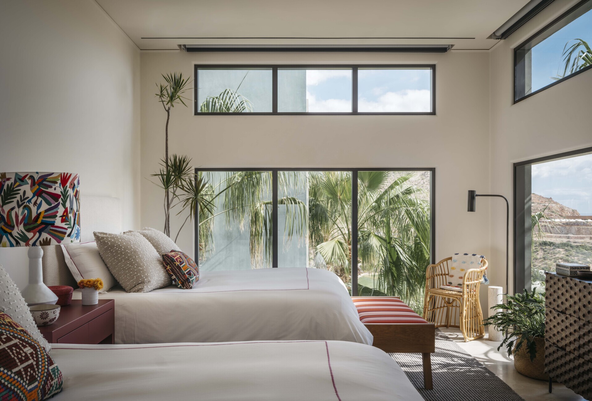 This bedroom feels like a little treehouse escape, nestled right into the palms. Tall windows on three sides pull in soft daylight and swaying greenery, giving the room a breezy, indoor-outdoor feel. The clean lines of the white bedding and simple bed frames keep things crisp, while colorful embroidered pillows and a vibrant lampshade add a playful, handmade touch. A mix of natural textures, wicker, wood, and a woven rug, brings a cozy, layered warmth that balances the room’s height and openness.
