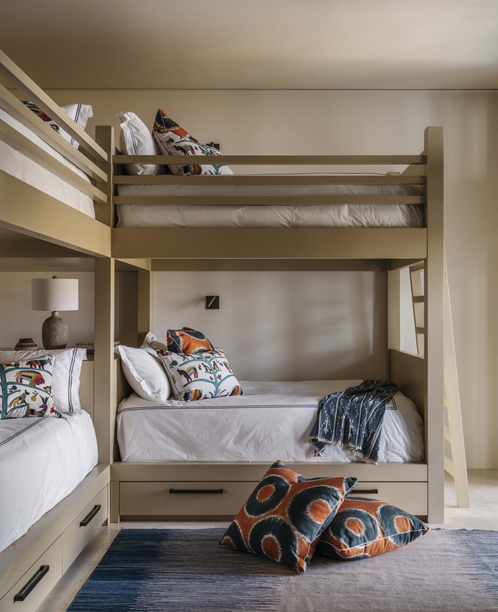 This bunk room strikes the perfect balance between playful and polished. The built-in beds, painted in a warm sandy beige, feel sturdy and thoughtfully designed, each with its own cozy nook and a drawer underneath for tucking things away. Crisp white bedding is brought to life with colorful embroidered pillows that hint at desert sunsets and handmade charm. A few bold ikat accents in burnt orange and deep navy add just the right amount of energy without overwhelming the calm. The soft ombré rug grounds the room and invites bare feet, while the scattered floor cushions suggest that this space was made for lounging, chatting, and a bit of mischief.
