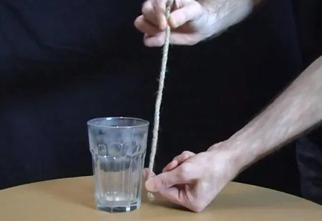 A hand holding a string above a glass of water on a table.