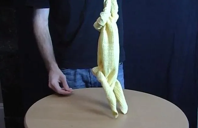 A person demonstrating a yellow cloth tied in a knot hanging from a table.