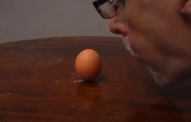 A person blowing air towards an orange egg on a wooden surface.