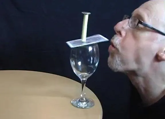 Man balancing a card on a glass with a nail on top.