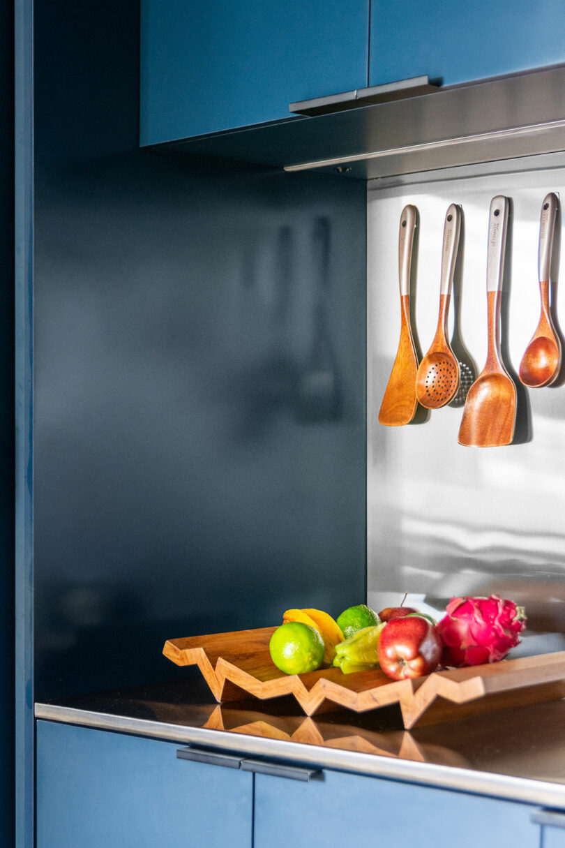 A modern kitchen with wooden utensils hanging on a rack and a geometric wooden tray holding various fruits on the counter.