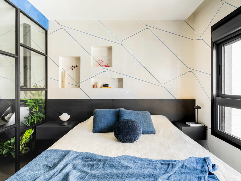 Modern bedroom with a bed against a geometric accent wall, built-in shelves, black framed glass partition, and large window letting in natural light.