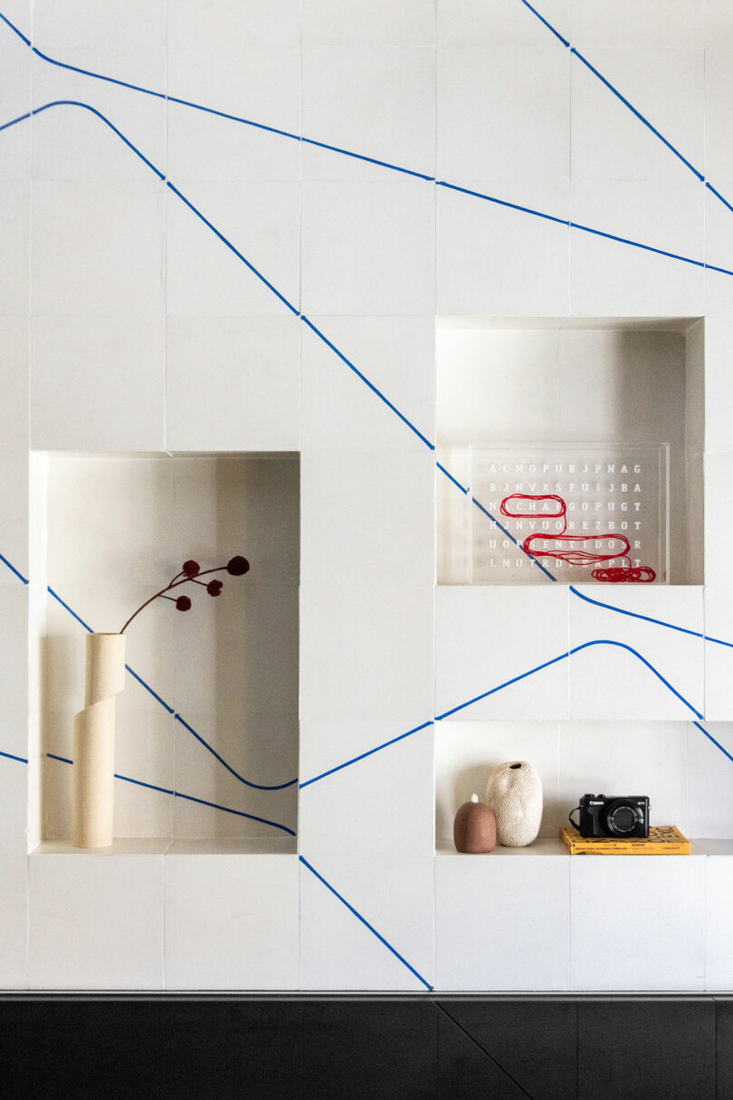 A white tiled wall with blue lines features recessed shelves holding a vase with red stems, a word clock, decorative vases, a camera, and yellow books.