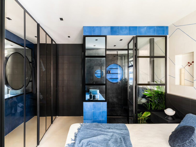 Modern bathroom with black and blue tiles, glass partitions, round mirrors, a small sink area, and a green plant next to a bed with a blue throw blanket.