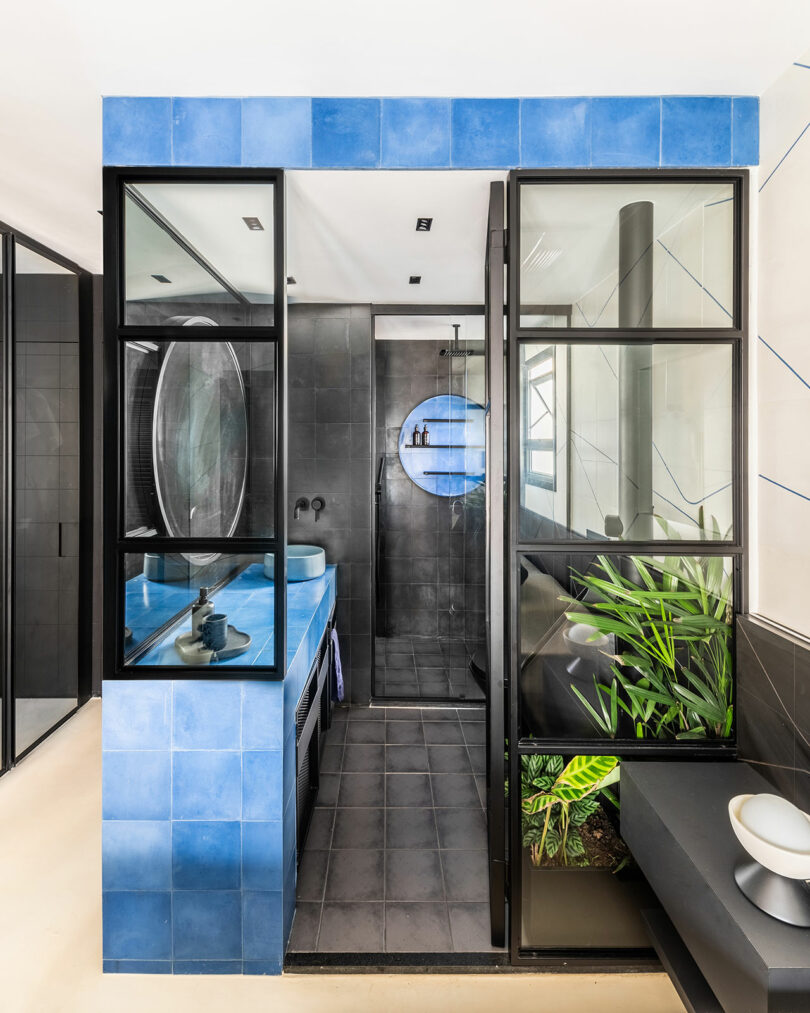 Modern bathroom with blue tiles, black-framed glass partitions, a black shower area, a rectangular sink, and green plants on a black countertop.