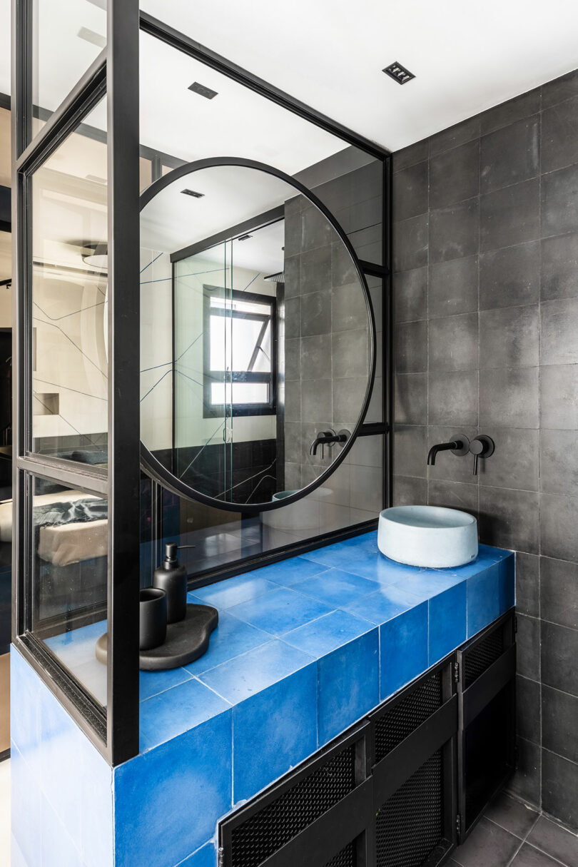 Modern bathroom with a large round mirror, blue countertop, light blue vessel sink, and matte black fixtures against dark tiled walls.