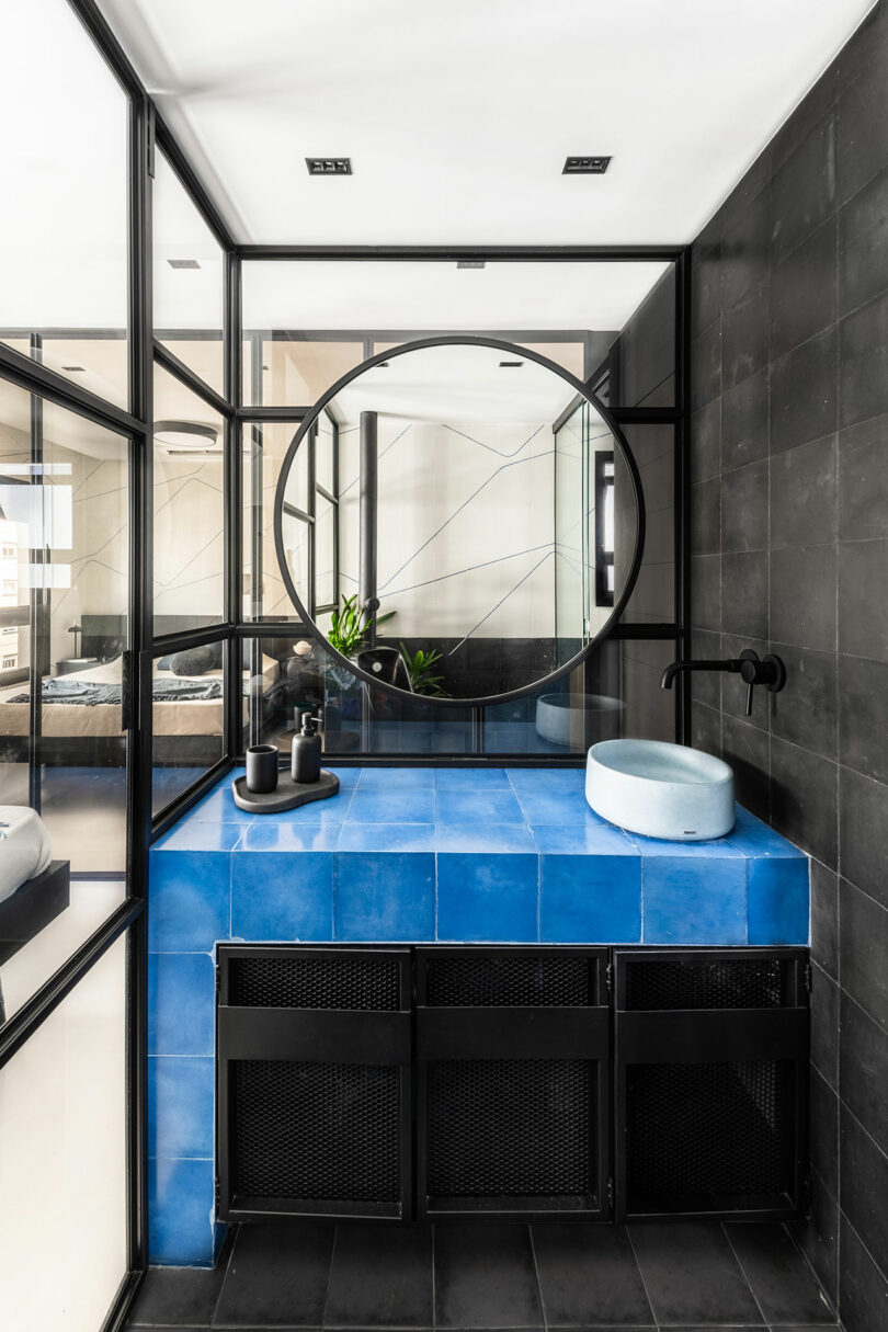 Modern bathroom with black tile walls, blue tiled vanity, round mirror, vessel sink, and glass partitions separating the space.