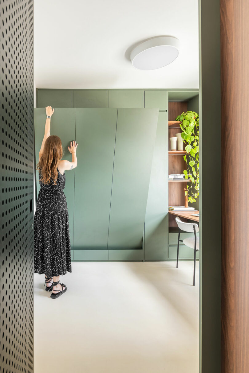 A person in a black dress opens a large, green built-in cabinet in a modern, minimal room with shelves, plants, and a chair.