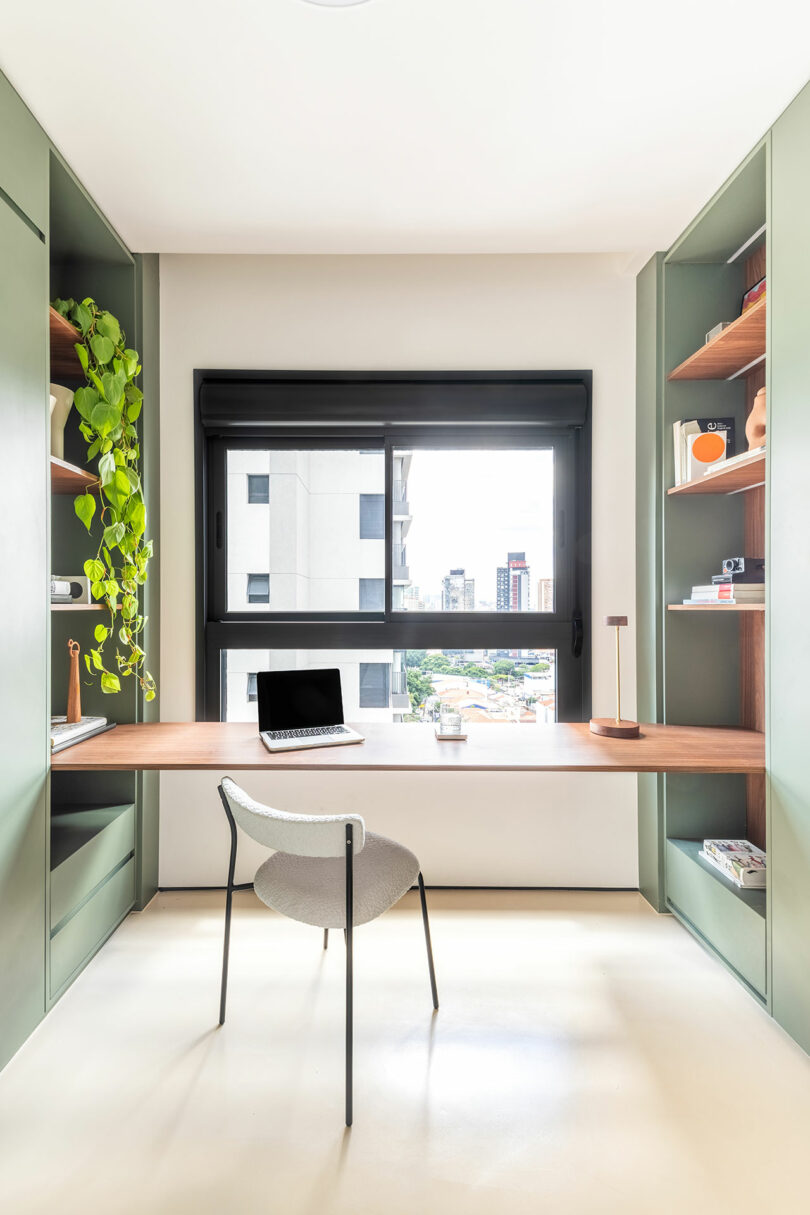 A modern home office with green built-in shelves, a wooden desk, a laptop, a chair, a potted plant, and a large window overlooking city buildings.