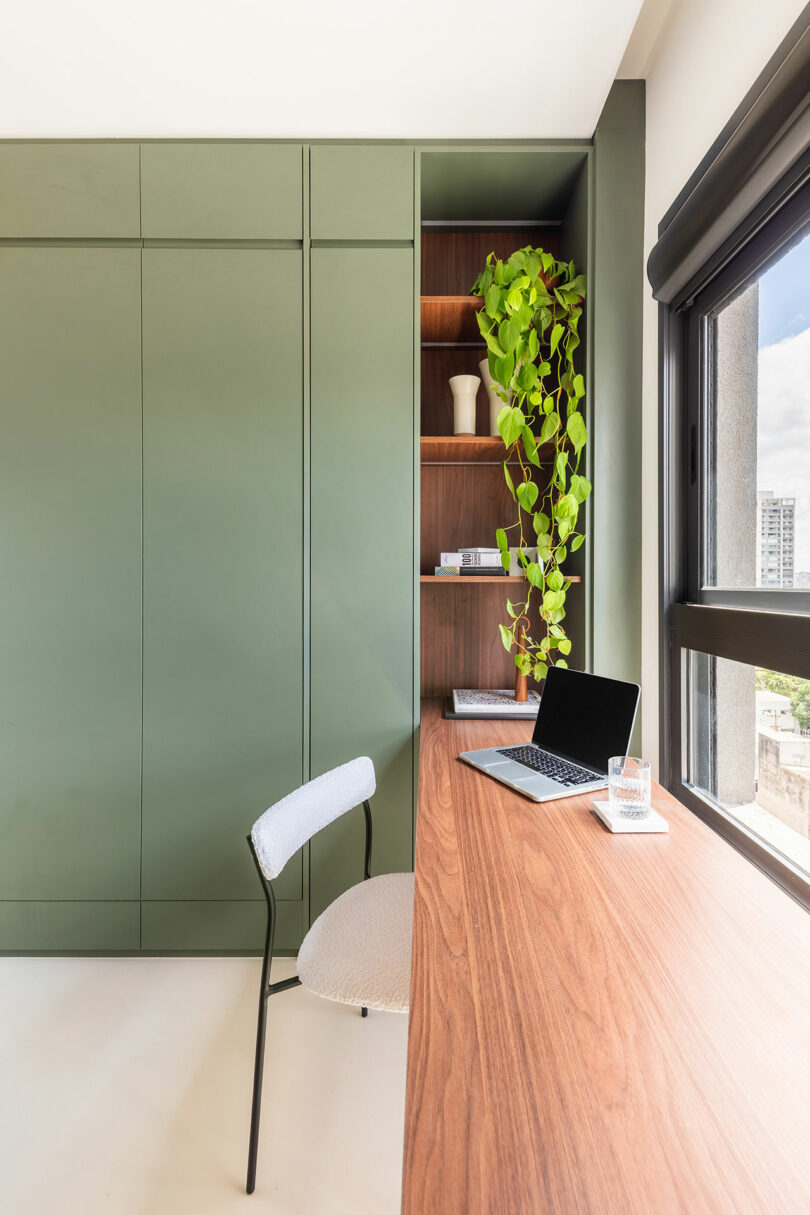 Minimalist home office with a wooden desk, green built-in shelves, a laptop, a chair, a potted trailing plant, and a large window letting in natural light.