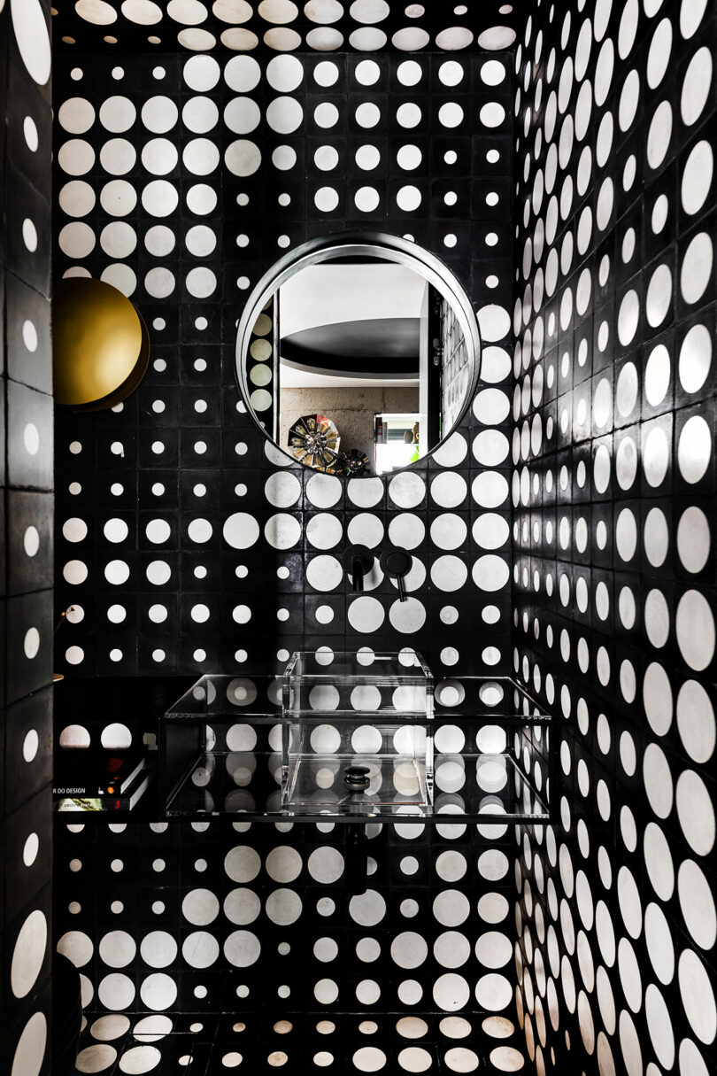 A small bathroom with black tiles covered in large white polka dots, a round mirror above a transparent sink, and a brass wall sconce.